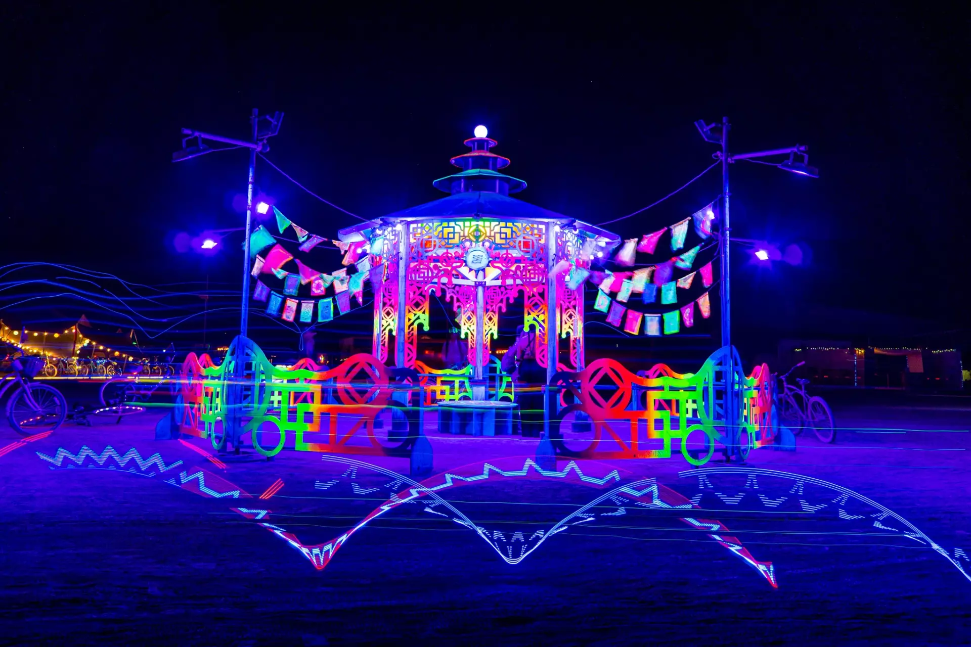 UV-lit temple art piece featuring prayer flags and laser cut structural elements illuminated at night on playa 