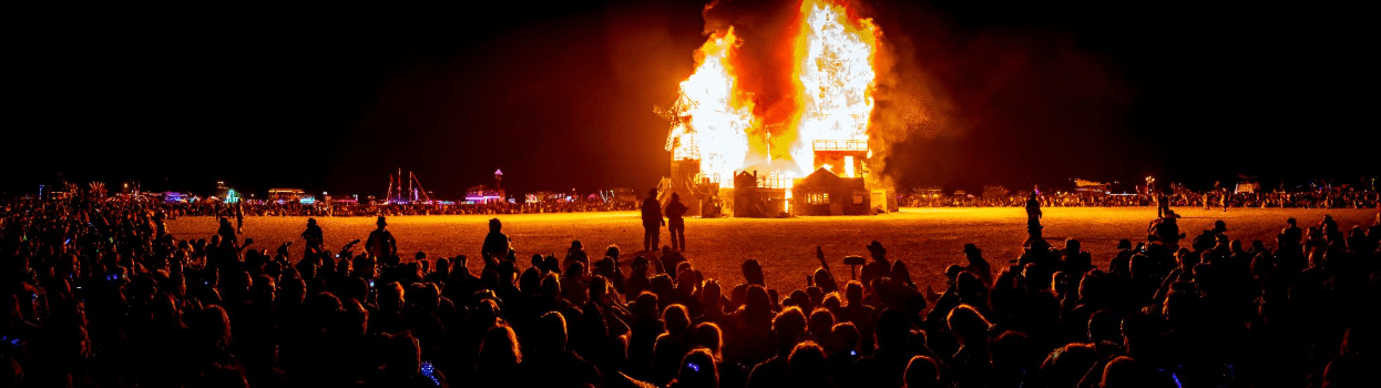 People seated in a large circle around a burning sculpture, with a few people standing between the crowd and the fire. 