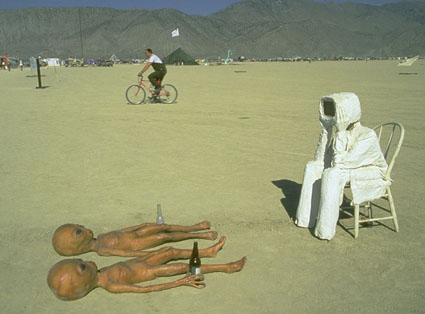 A figure in a white astronaut suit sits on a white chair, elbows on knees and hands on chin, looking at two alien figures holding beer bottles who are lying on the playa.