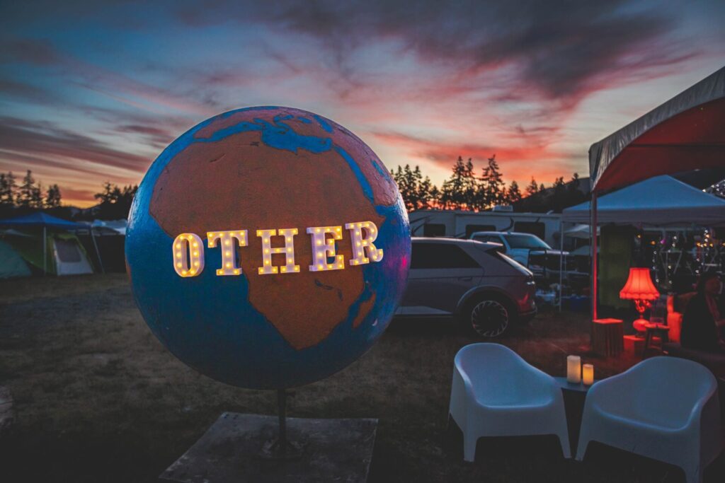 A large globe with the word "OTHER" in circus lights at sunset in front of a forest.