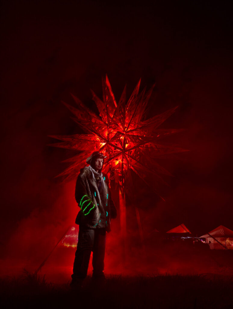 A man in an coat embellished with LED details stands in front of a spiky wooden effigy on playa that is beginning to burn.