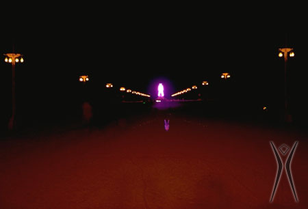 The Man effigy lit with red lights seen at a distance at night. Lanterns line the street to the Man, casting a red glow on the playa.