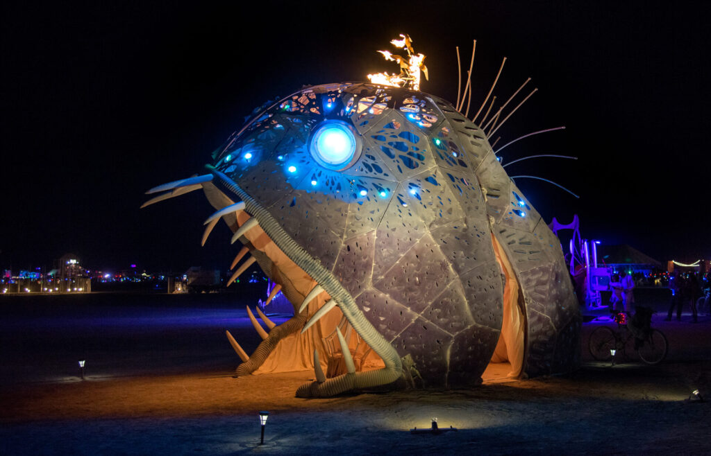 An angler fish with open mouth showing pointy teeth waits for people to walk inside. 