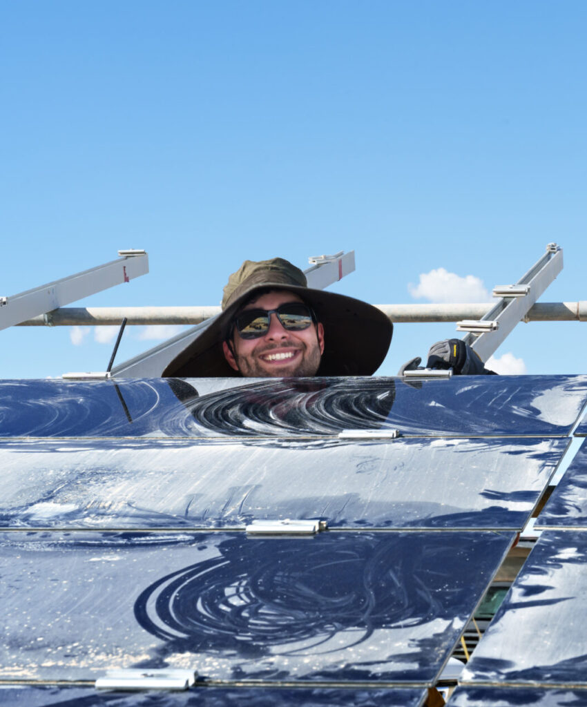 A smiling white man wearing sunglasses and a hat with a large brim peeks over a shiny black solar panel.