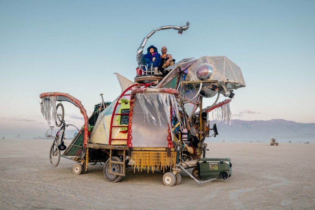 Two burners take in the sunrise atop a mutant vehicle resembling a silver turtle. 