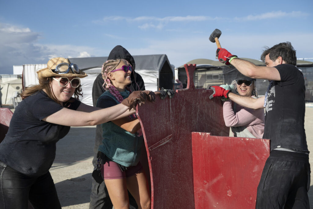 Volunteers help set up Videogasm camp. Four women constructing something for their camp with red painted wood. One woman has a hammer. 