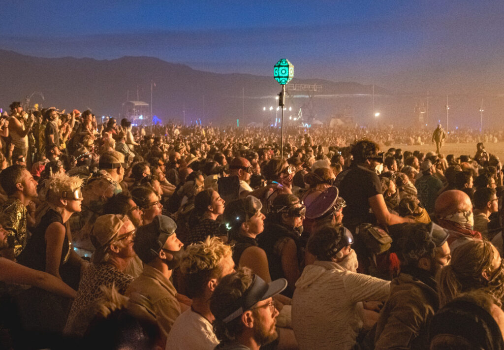A crowd of people in the glow of firelight, looking away from the camera