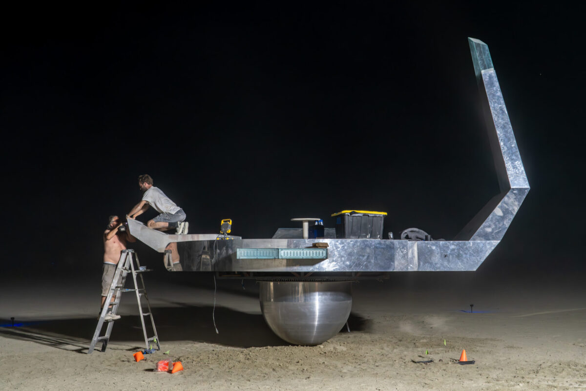 A person on a ladder hands parts to a person balanced on a shiny metal platform balanced on a sphere.