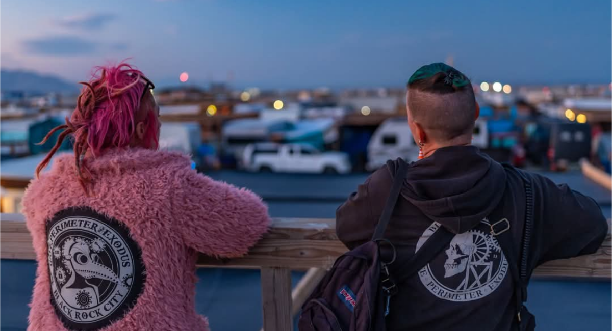 At sunrise, two people wearing hoodies emblazoned with "Gate, Perimeter, Exodus" emblems stand on a wood balcony overlooking Black Rock City.
