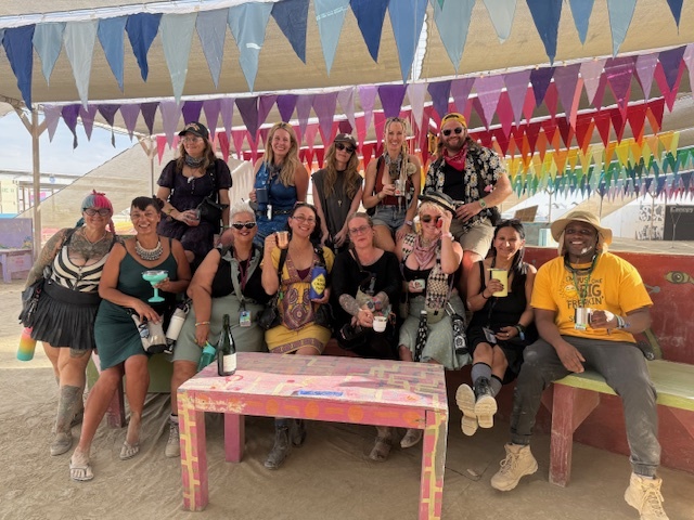 Thirteen smiling people sit under a shade structure decorated with rows of colorful pennants  hung from the ceiling.