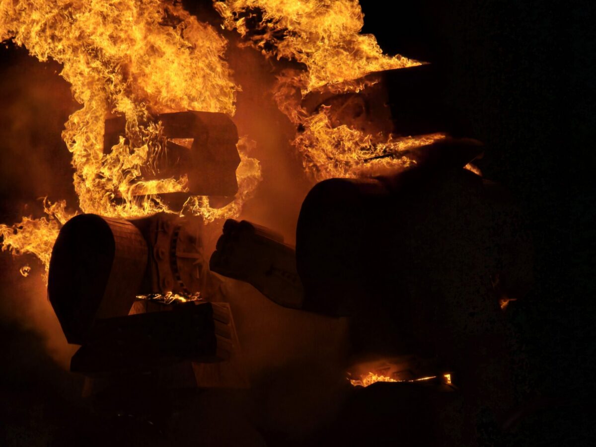 Close up of two wooden figures evocative of the rock-em-sock-em-robots game, on fire. 